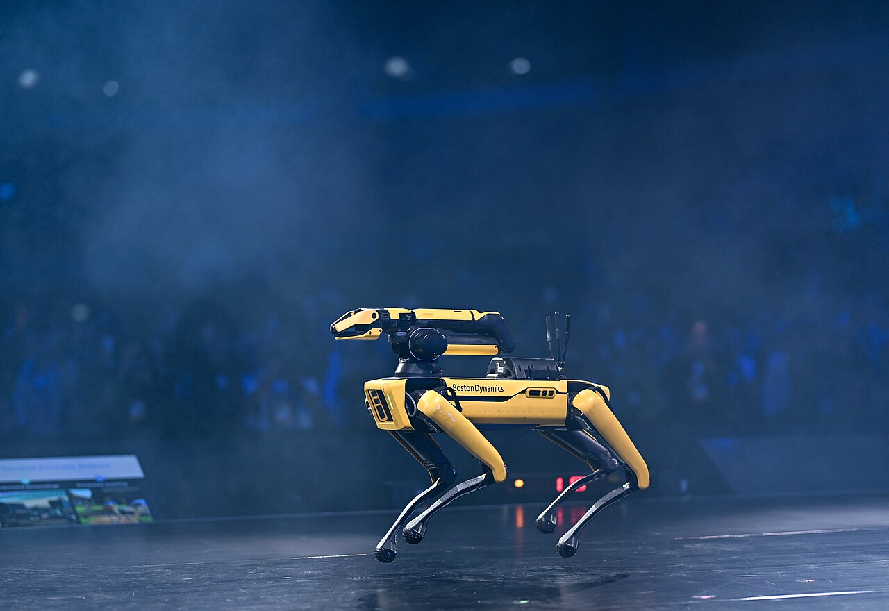 A yellow walking robot with an articulated arm, mid-stride on a blue stage
